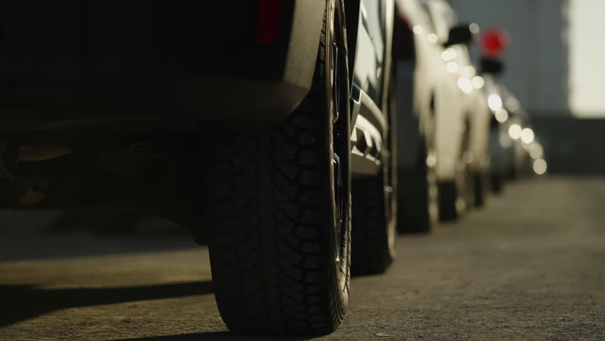 Low-angle close-up of a row of parked vehicles focusing on rugged tires and alloy wheels, bathed in golden sunlight along an urban street with textured asphalt and long shadows