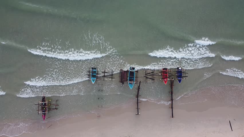 Aerial top view of colorful local fishing boat docked at the sandy beach with ocean wave crashing on the shore.