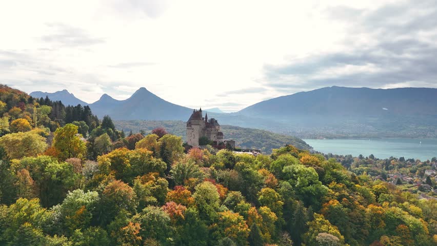 Medieval Château de Menthon‑Saint‑Bernard perched on wooded hillside, revealing its towers and turrets framed by colourful fall foliage, tLake Annecy and layers of Alpine mountains, drone slow push in