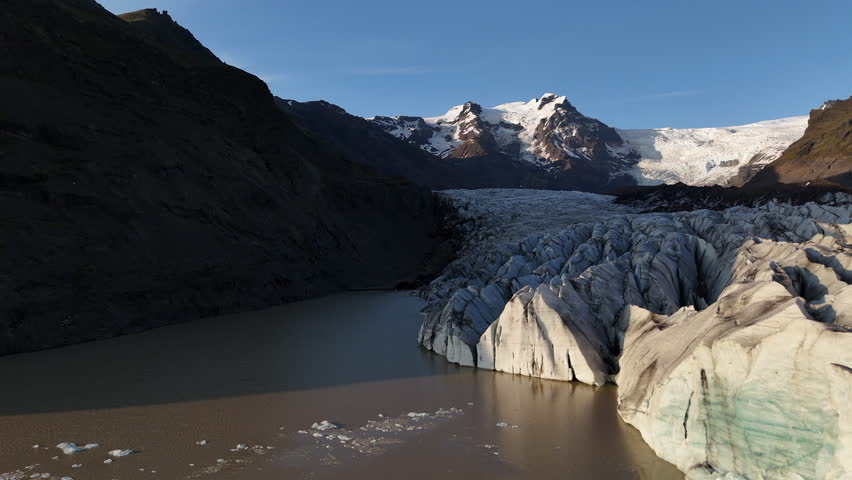 Aerial view of Svínafellsjökull Glacier at sunset, showing deep ice crevasses flowing into a dark glacial lagoon, framed by steep volcanic mountains and warm evening light in southern Iceland.
