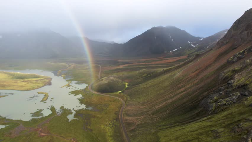 An aerial wide-angle shot captures a dramatic and vast mountain landscape under a cloudy sky. A brilliant rainbow arcs prominently over the scene, its colorful light illuminating a portion of the valley below on the left. The valley floor features a large, winding river or body of water surrounded by lush green and yellow tundra-like vegetation. Steep mountain slopes rise on the right side and in the background, showing a mix of earthy tones and patches of green moss and lichen. A narrow, winding dirt road cuts through the valley floor, adding a sense of scale and remoteness to the scene. The misty clouds and soft light enhance the overall atmospheric quality of this pristine natural environment.