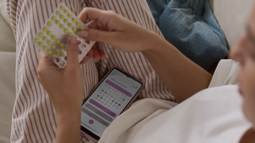 Over-the-shoulder shot of young Caucasian woman taking her medication while lying in relaxed position on couch with period calendar opened on her smartphone
