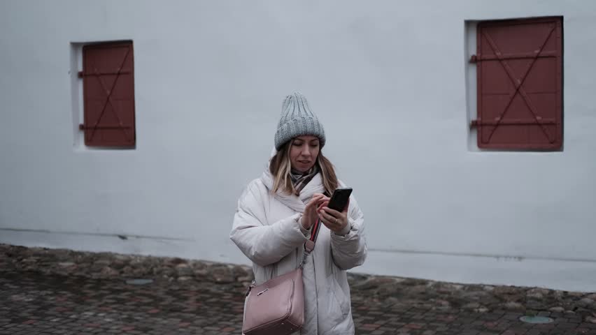 Portrait of mature woman in wool hat standing on the urban street with mobile phone in hand. Middle-aged 40s female using smartphone, walking the city at cold autumn day. Living in modern district