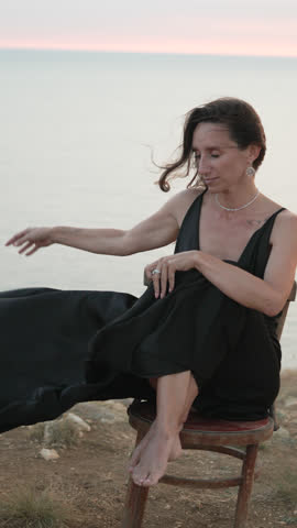 Woman, nature, dress, thoughtful woman in black dress sits on chair by ocean, hair blowing in wind, vertical video
