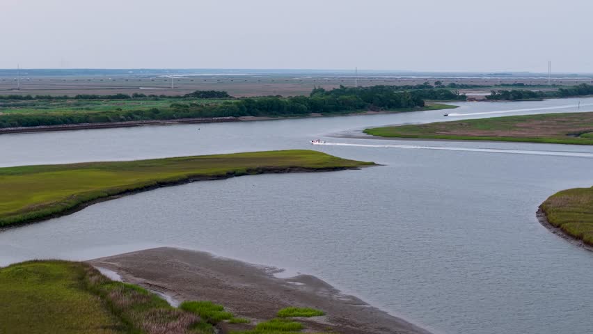 Aerial drone view of a winding river cutting through green marshland near Charleston, South Carolina, as a boat travels along the waterway under a blue sky. Serene lowcountry landscape.