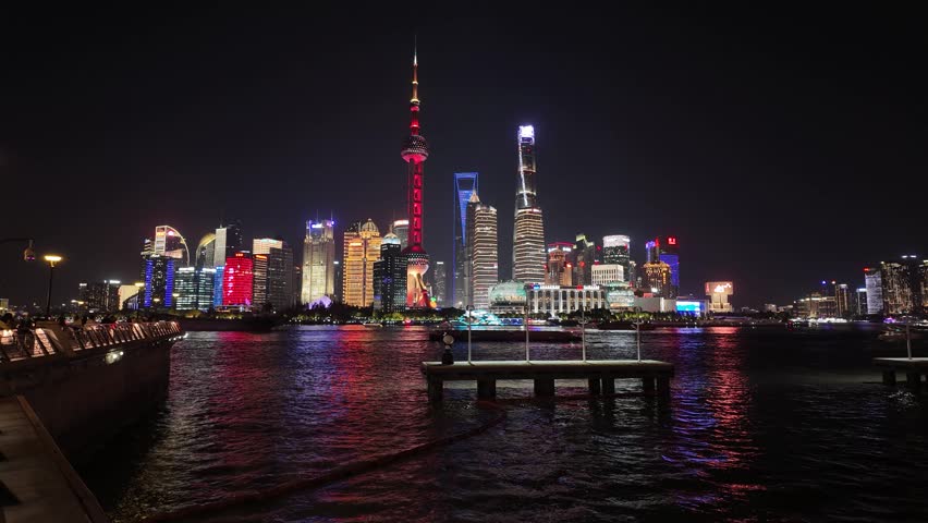 Night timelapse of iconic Shanghai Pudong skyline with Oriental Pearl Tower and Shanghai Tower reflecting on calm Huangpu River, colourful lights