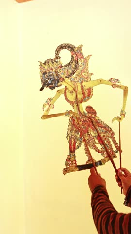 Puppeteer's hands manipulating a traditional Indonesian Wayang Kulit leather puppet during a performance, telling an ancient epic story with intricate movements against a warm, light background