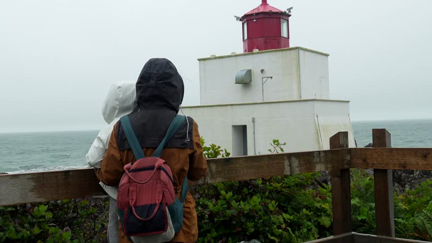 Tourists wearing raincoats and backpacks pointing at the ocean near amphitrite point lighthouse in ucluelet, vancouver island, british columbia, canada, on a cloudy day