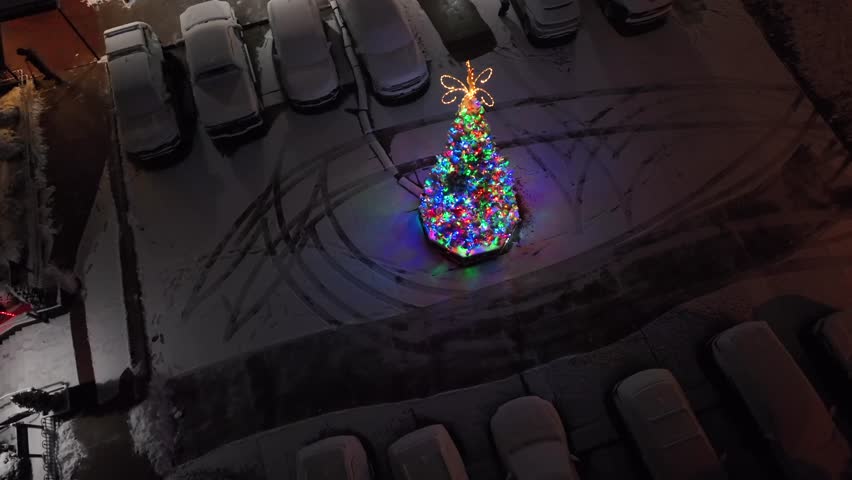 Aerial top down of Colorful Christmas tree glowing in snowy parking lot, cars covered in fresh snow, vibrant holiday lights reflecting across cold night. Festive winter scene in American town.