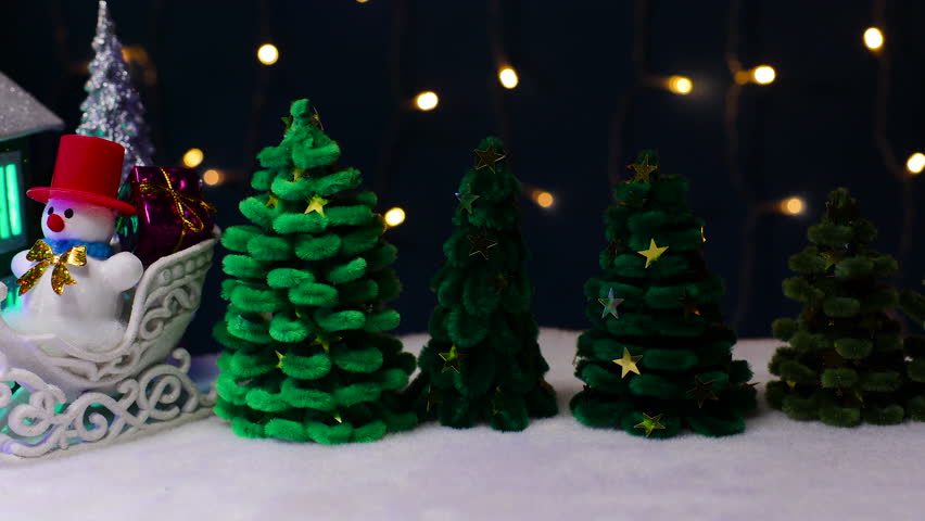 On a blue background with flashing lights, there are many green Christmas trees and a two-color house with letters and a snowman on a sled