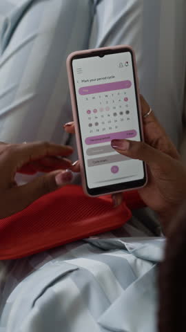 Vertical of young Black woman lying on sofa with heating pad on stomach while checking cycle calendar on smartphone in quiet home setting