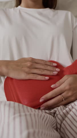 Vertical cropped shot showing unrecognizable woman seated on couch holding heating pad on abdomen to soothe menstrual pain