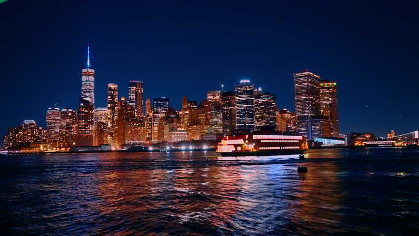 New York City skyline lights up at night while a ferry moves across the water. This scene shows the buildings reflecting on the river, creating a vibrant atmosphere.