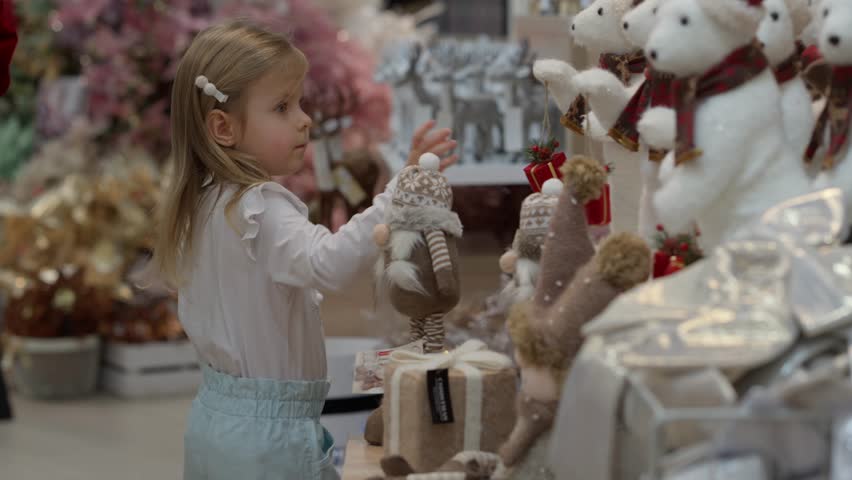 A charming child looks at toys in a store. A cute little girl in a store among Christmas decorations. A child girl chooses toys in a store. A child plays with Christmas teddy bears.