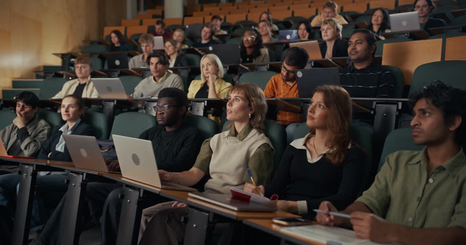 Diverse Group of University Students Sit in Lecture Theater, Listening, Typing on Laptops, and Writing Notes During Academic Lecture, Higher Education Learning