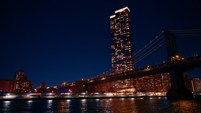Bright lights shine from buildings in New York City at night. A large bridge connects the structures, reflecting on the water below. The scene captures urban life and architecture.