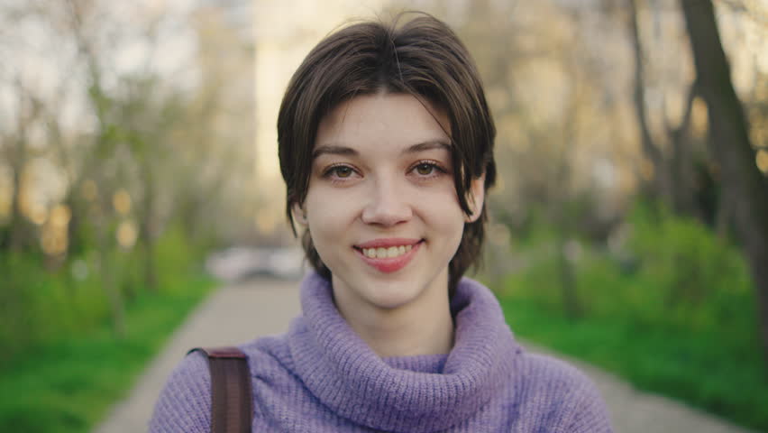Young woman stands on path in green park setting during daytime with city buildings visible in background, smiling softly and crossing arms