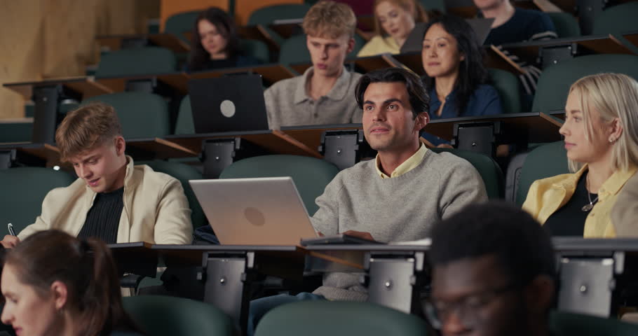 In University Lecture Hall, a Male Student Surrounded by Diverse Classmates Listens Intently, Raises His Hand, and Confidently Answers the Professor’s Question, Using a Laptop