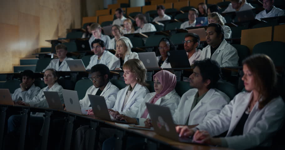 Diverse Group of University Science Students in White Lab Coats Absorb a Complex Lecture, Typing on Laptops and Taking Notes Auditorium