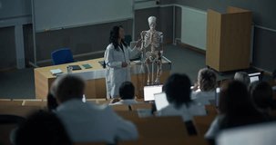 Asian Female Science Professor in White Lab Coat Leads University Anatomy Lecture, Using a Detailed Skeleton While Diverse Medical Students use Laptops in Classroom, Clinical Education and Teamwork. - Powered by Shutterstock - Get 15% off with code: PIKWIZARD15