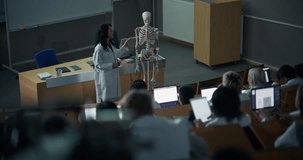Science Professor in a White Lab Coat Delivers an Interactive Anatomy Lecture, Teaching Skeletal Structure on a Model While Diverse University Students in Lab Coats Follow Digital Notes on Laptops - Powered by Shutterstock - Get 15% off with code: PIKWIZARD15