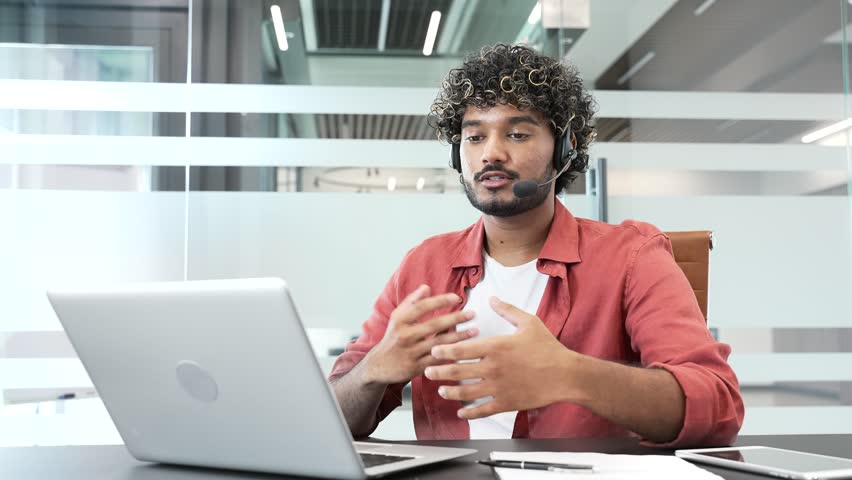 Young handsome businessman in a headset talking on a video call using laptop computer sitting at workplace in office. Entrepreneur has a business meeting. Manager speaks remotely at online conference