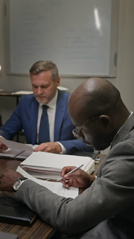 Medium vertical shot of African American and Caucasian male attorneys discussing clause in legal document, while inspecting case evidence, preparing arguments for court litigation