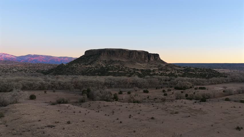 Wide shot of the flat-topped Black Mesa rising from the arid desert floor. The scene captures the high-altitude Southwestern landscape under the soft, receding light of the golden hour.