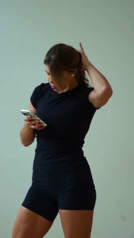 Vertical shot of a woman in black sportswear standing indoors, looking down intensely at her smartphone held in her hands. She is taking a break from exercise or checking social media messages