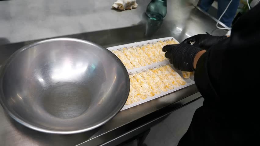 A professional kitchen worker wearing black nitrile gloves places breaded shrimp into a metal bowl before submerging them in a deep fryer to cook until they are golden brown and crispy.