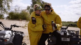 Couple in yellow raincoats on beach sharing drink beside parked quad, goggles and cans in hand, sandy shore and sunlit trees in background, playful laughter - Powered by Shutterstock - Get 15% off with code: PIKWIZARD15