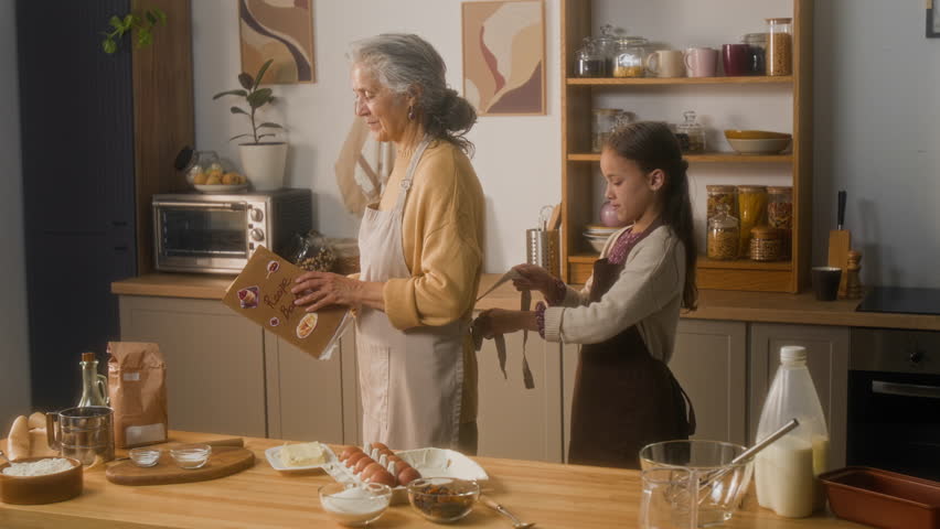 Medium long shot of young Caucasian girl standing next to mature grandmother with recipe book while choosing what to cook at home kitchen