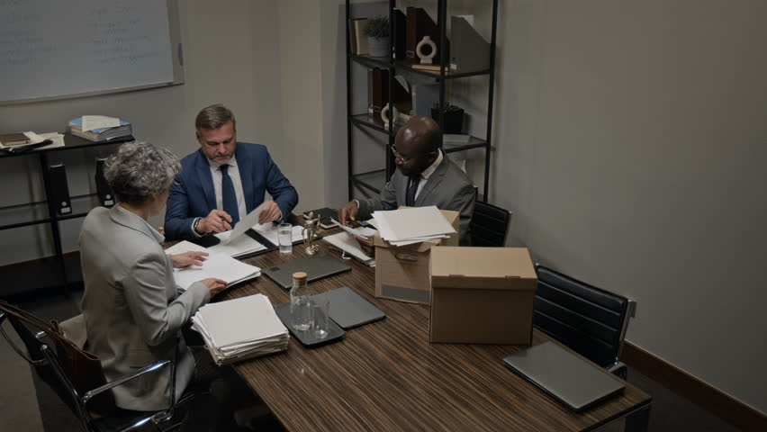 Wide high-angle security camera shot of multiethnic team of defense attorneys sitting around table in meeting room, researching paperwork for precedents while preparing for lawsuit