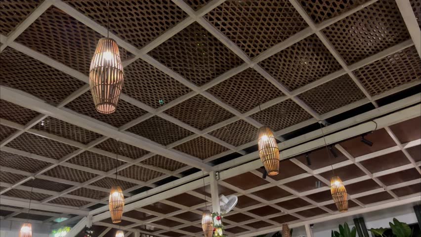 Smooth panning shot of multiple lantern‑shaped bamboo pendant lamps hanging from a white ceiling grid with woven wood panels