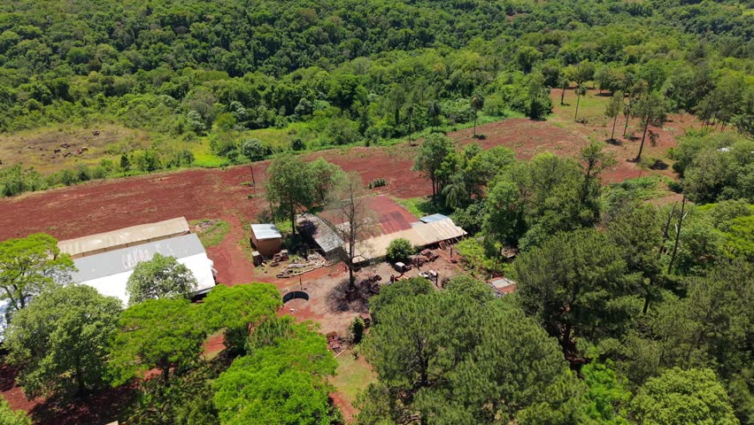 Aerial view reveals a small, rustic farmstead nestled against a dense green forest, surrounded by bright red earth fields typical of Misiones, Argentina.