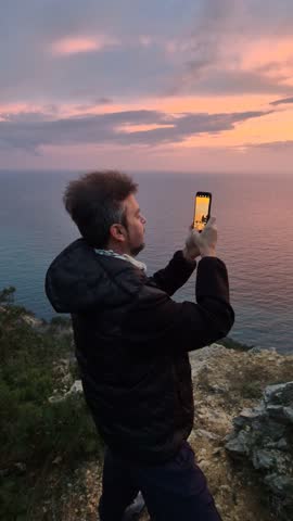 Smartphone sunset cliff, man capturing dramatic ocean view with mobile phone during twilight.