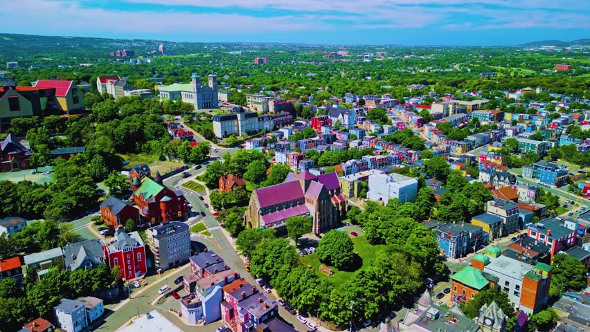 A summer day in downtown St. John’s captured from above, showcasing the iconic jelly bean row houses in vibrant colors lining the streets. The aerial perspective highlights the city’s unique charm, wi