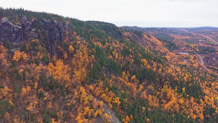 Rural valley features a curving road with small houses and farms among brilliant orange and green trees; a river and distant mountains frame the scene.