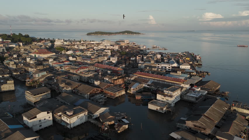Gentle aerial view of the bustling downtown port and dense coastal city of Tanjung Pinang at sunrise. Buildings built over the water, small docks, and a soft golden light. Tanjungpinang, Indonesia.