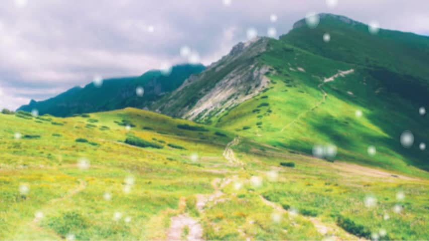 "Green mountain ridge with a winding hiking trail under a cloudy sky with a bokeh snow overlay."