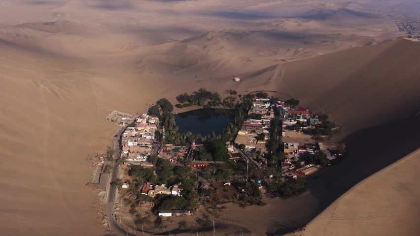 Winding dune slopes fall toward the oasis village creating layered depth across the sandy basin, aerial closeup descend