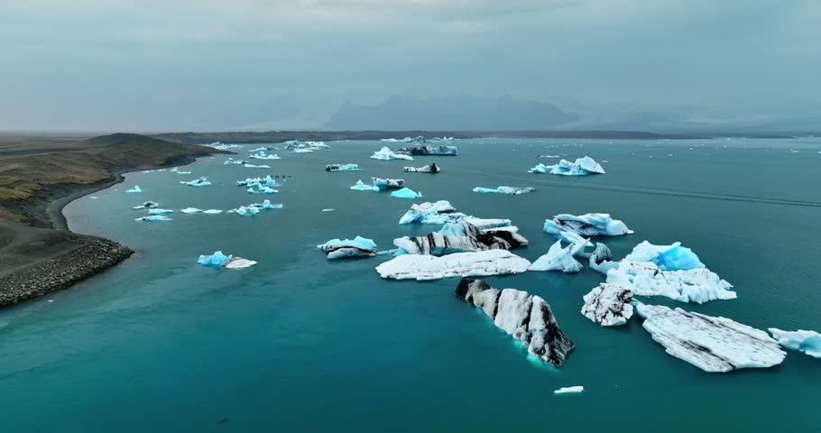 A smooth aerial orbit reveals drifting Icelandic icebergs scattered across a wide lagoon, with layered blue surfaces, dark streaks, and a rugged shoreline under a cold Arctic sky.
