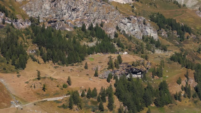 Aerial panning right revealing an isolated rock cabin in a pine forest in Italy, Aosta Valley, Valle d’Aosta, surrounded by alpine mountains