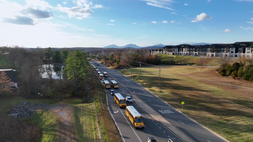 Aerial view of long row yellow school bus moving through America suburb under fall sun, lined with trees, pond reflection and modern apartment complex. blue mountain range in distance.