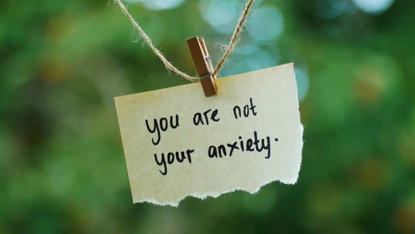 Close-up inspirational note reading "you are not your anxiety" pinned on twine, isolated against a bright natural green bokeh background using soft daylight.