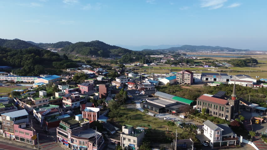 4K UHD drone aerial footage of a peaceful countryside village in South Korea. The shot shows colorful rooftops, rural roads, farmland patterns and green hills stretching toward the horizon under a cl