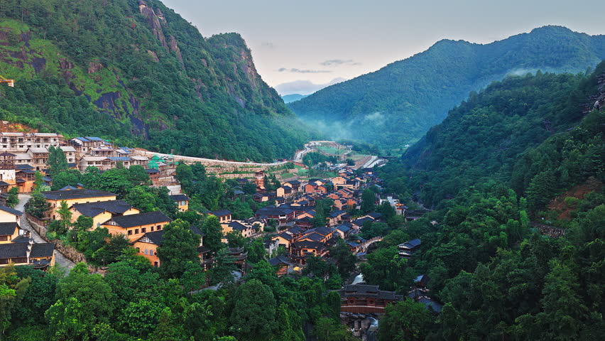 Aerial shot of traditional ancient village with classic architecture in a lush green mountain valley at dusk. The famous Wangxiang Valley Scenic Area is located in Jiangxi, China.