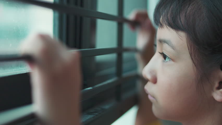Close up side profile of sad Asian girl looking out window with selective focus. Lonely child face details with blurred hand holding bars in foreground, concept of waiting, depression, or boredom.