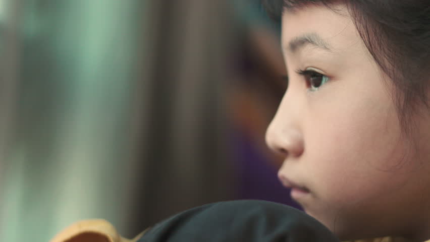 Close up side profile of sad Asian girl face looking away with selective focus. Lonely child sitting near window feeling bored, depressed, unhappy, thinking or waiting in solitude at home.