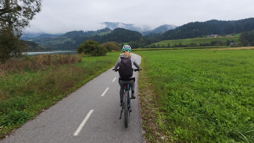 Young woman rides an e-bike along a peaceful countryside path near the Drava River, surrounded by open fields and distant hills. Ideal for outdoor lifestyle, fitness, travel and adventure themes
